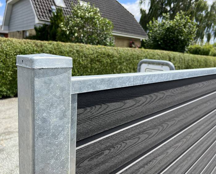 Nærbillede af en Komposit Hegn med galvaniseret bundplader, stålstolper & u-skinner – Type 2, med vandrette trælignende paneler indrammet af en galvaniseret metalstolpe og topskinne. Hegnet er placeret udendørs med en pænt klippet hæk og et hus med tegltag i baggrunden under en solrig himmel.