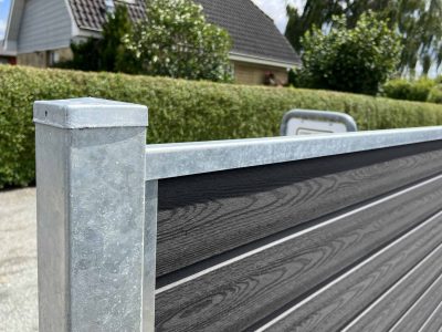 Nærbillede af en Komposit Hegn med galvaniseret bundplader, stålstolper & u-skinner – Type 2, med vandrette trælignende paneler indrammet af en galvaniseret metalstolpe og topskinne. Hegnet er placeret udendørs med en pænt klippet hæk og et hus med tegltag i baggrunden under en solrig himmel.