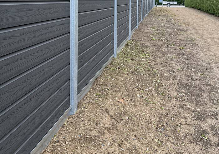 Udsigt over en grus- og jordsti langs en lang Type 2 komposit hegn med galvaniseret bundplader, stålstolper & u-skinner. Spredte blade og små småsten dækker jorden. Hegnet strækker sig mod baggrunden, hvor et hvidt køretøj er delvist synligt.