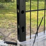 Et nærbillede af en dobbeltlåge 2D 6/5/6 mm Sort RAL 9005 Komplet Inkl. stolper, der viser den sorte metalstolpe med laserskåret forboret skrog, fastgjort til et ståltrådspanel med en sort kabelbinder og plastfolie. Græs og træer ses i dagslysbaggrunden.