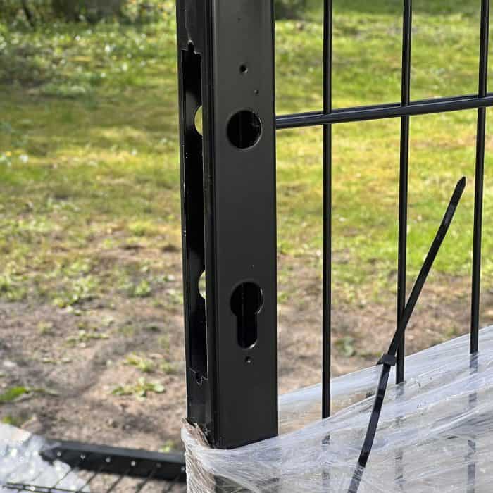 Et nærbillede af en dobbeltlåge 2D 6/5/6 mm Sort RAL 9005 Komplet Inkl. stolper, der viser den sorte metalstolpe med laserskåret forboret skrog, fastgjort til et ståltrådspanel med en sort kabelbinder og plastfolie. Græs og træer ses i dagslysbaggrunden.