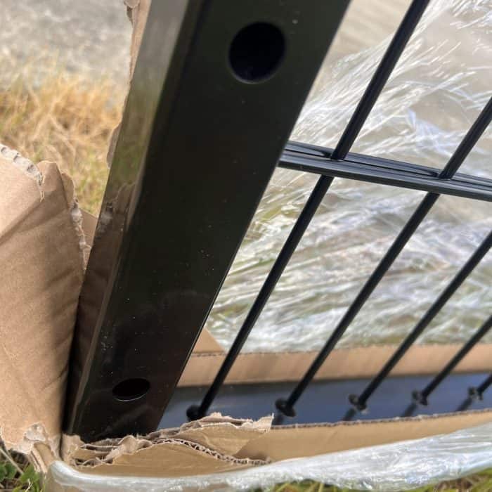 Et nærbillede af en Dobbeltlåge 2D 6/5/6 mm Sort RAL 9005 Komplet Inkl. stolper hegnspæl og trådpanel, med laserskåret skrog, delvist pakket ind i plastik inde i en iturevet papkasse, græs og fortov synligt i baggrunden.