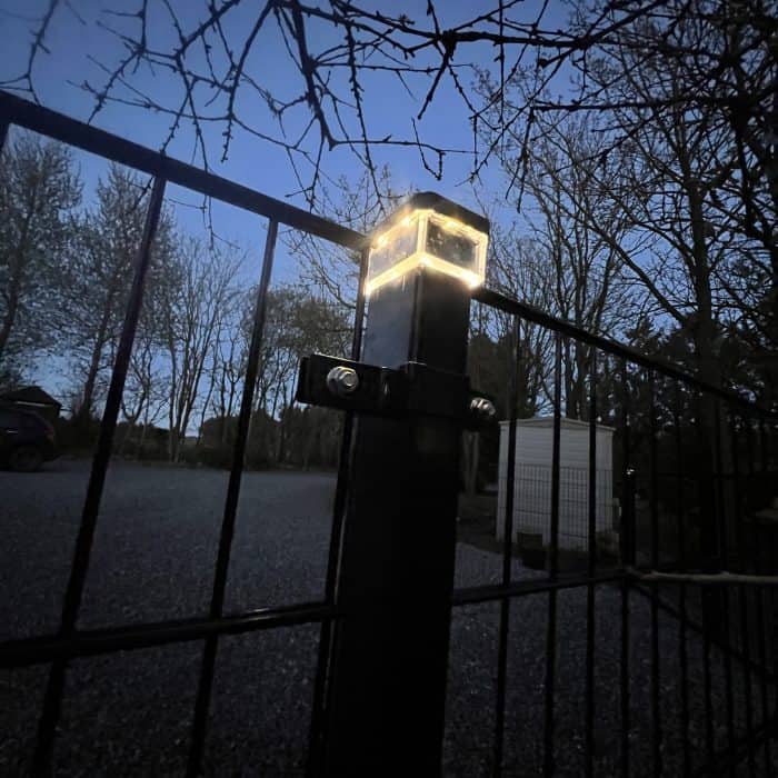 En stålstolpe med Mellemstolpe Beslag 60x40mm til Panelhegn Sort RAL 9005 (Designserie) og et lille, oplyst lys på toppen står foran nøgne trægrene i skumringen, med en grusbelagt indkørsel, en parkeret bil og et skur i baggrunden.