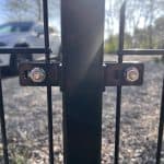 Nærbillede af en sort Mellemstolpe Beslag 60x40mm til Panelhegn Sort RAL 9005 (Designserie) fastgjort med to bolte til en stolpe. En sløret hvid bil og solbeskinnede træer ses i baggrunden.