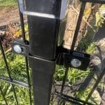 Nærbillede af en Mellemstolpe Beslag 60x40mm til Panelhegn Sort RAL 9005 (Designserie) med beslag og bolte, der fastgør den til 2D Panelhegn, omgivet af grønne planter og udendørs vegetation i sollys.