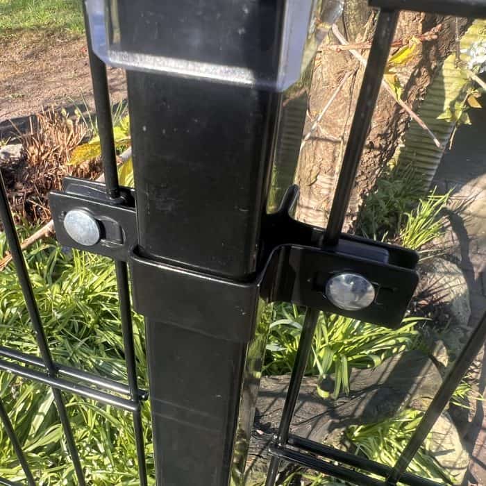 Nærbillede af en Mellemstolpe Beslag 60x40mm til Panelhegn Sort RAL 9005 (Designserie) med beslag og bolte, der fastgør den til 2D Panelhegn, omgivet af grønne planter og udendørs vegetation i sollys.