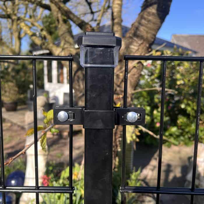 Et nærbillede af en sort 2D panelhegn fastgjort til en stålstolpe ved hjælp af Mellemstolpe Beslag 60x40mm til Panelhegn Sort RAL 9005 (Designserie). Træer, planter og en del af et hus fremstår sløret i den solrige baggrund.