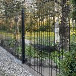 Et sort 2D Panelhegn med Mellemstolpe Beslag 60x40mm til Panelhegn Sort RAL 9005 (Designserie) står ved en frodig have med grønne planter, sten og blomster. Sollyset gennem træerne kaster små skygger på græsset.