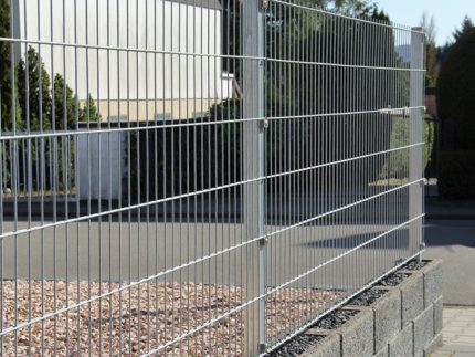 A Panelhegn 2D 6/5/6 mm HDG Galvaniseret - Komplet Inkl. Stolper & Beslag er monteret på en lav murstensmur, der omgiver et område med små sten. Det går parallelt med en asfalteret vej; træer og en bygning ses i baggrunden under en klar himmel.