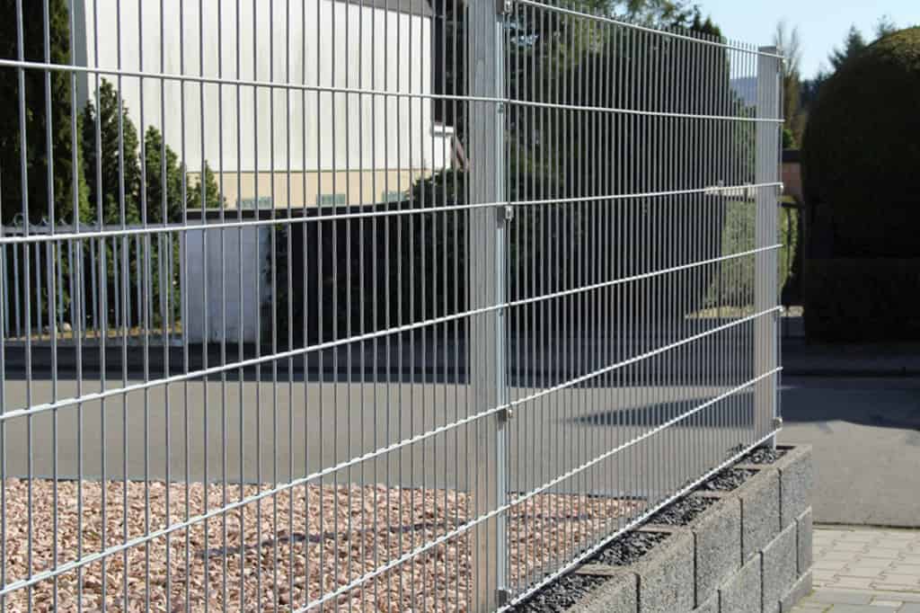 A Panelhegn 2D 6/5/6 mm HDG Galvaniseret - Komplet Inkl. Stolper & Beslag er monteret på en lav murstensmur, der omgiver et område med små sten. Det går parallelt med en asfalteret vej; træer og en bygning ses i baggrunden under en klar himmel.