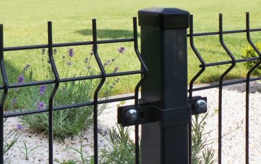 Nærbillede af et sort metalhegn med lodrette og bølgede vandrette stænger, med en Endestolpe Beslag 60x40mm til Panelhegn Sort RAL 9005 (Designserie) i midten. Bag hegnet er der en anlagt have med hvide småsten, lavendelplanter og et grønt græsareal i baggrunden. Sollys oplyser scenen.