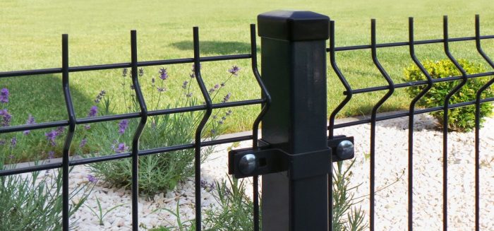 Nærbillede af et sort metalhegn med lodrette og bølgede vandrette stænger, med en Endestolpe Beslag 60x40mm til Panelhegn Sort RAL 9005 (Designserie) i midten. Bag hegnet er der en anlagt have med hvide småsten, lavendelplanter og et grønt græsareal i baggrunden. Sollys oplyser scenen.