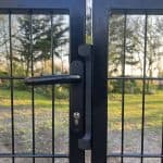 Et nærbillede af Dobbeltlåge 2D 6/5/6 mm Sort RAL 9005 Komplet Inkl. stolper metalporten med håndtag og lås, placeret udendørs med træer og græs i baggrunden, mens sollys filtreres igennem.