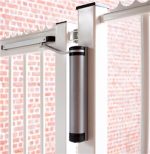 En moderne LION Kompakt Hydraulisk Portlukker fra Locinox er monteret på en hvid metalport. Den tættere er cylindrisk med en slank sølv krop og sorte accenter i enderne. Baggrunden har en rød murstensvæg.