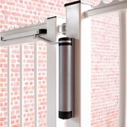 En moderne LION Kompakt Hydraulisk Portlukker fra Locinox er monteret på en hvid metalport. Den tættere er cylindrisk med en slank sølv krop og sorte accenter i enderne. Baggrunden har en rød murstensvæg.