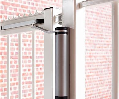 En moderne LION Kompakt Hydraulisk Portlukker fra Locinox er monteret på en hvid metalport. Den tættere er cylindrisk med en slank sølv krop og sorte accenter i enderne. Baggrunden har en rød murstensvæg.