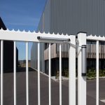 En hvid metalport med lodrette stænger og en Kompakt Hydraulisk Portlukker "LION" fra Locinox er delvist lukket foran en moderne industribygning med korrugeret metalbeklædning og træaccenter under en klar blå himmel.