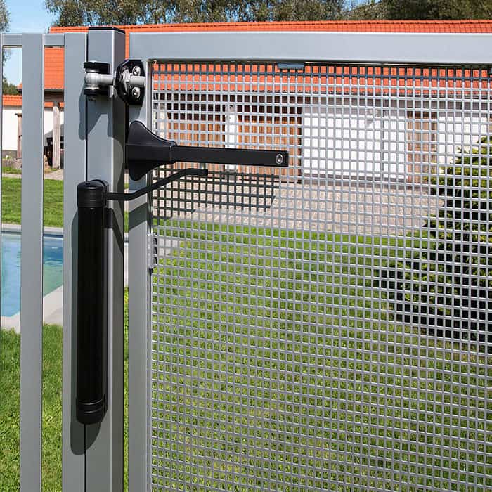 Et nærbillede viser en metallåge med mesh-design og glat automatisk lukning. Med Kompakt Hydraulisk Portlukker "LION" fra Locinox i slank sort, er den sat mod græs, en glitrende pool og bygninger med levende orange tage.