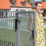 En person i en gul og hvid kjole står ved en åben Locinox Kompakt Hydraulisk Portlukker "LION." Bagved er et hus med orange tage og en pool. Personen smiler og nyder den solrige dag.