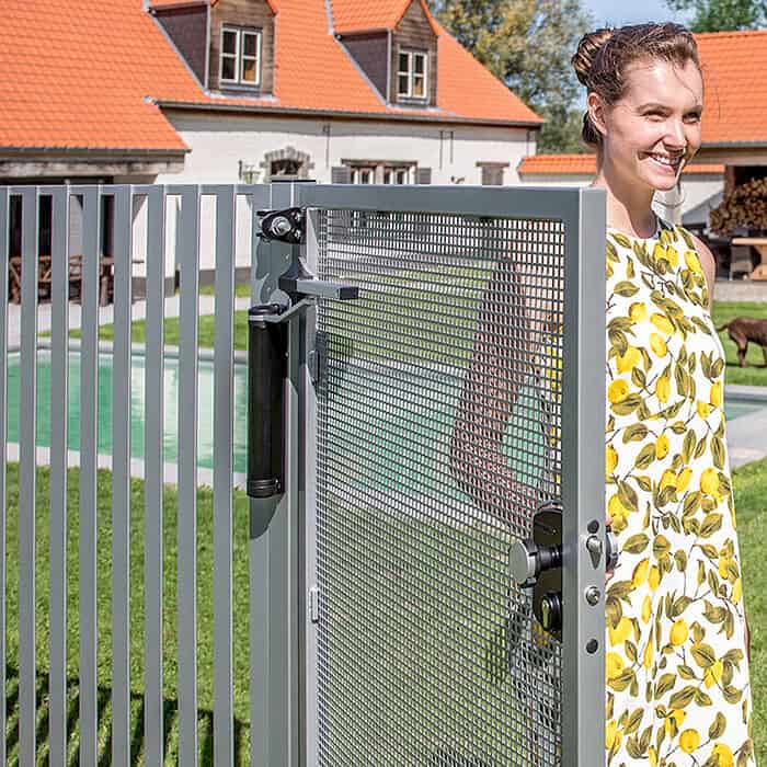 En person i en gul og hvid kjole står ved en åben Locinox Kompakt Hydraulisk Portlukker "LION." Bagved er et hus med orange tage og en pool. Personen smiler og nyder den solrige dag.