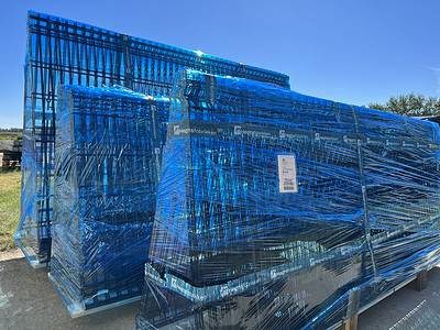 Stabler af store blå paller pakket ind i klar plast er placeret udendørs under en lys, klar himmel. Ved at løfte byrderne på træbaser pakkes pallerne sikkert sammen med nogle etiketter og skilte synlige gennem plastfolien.