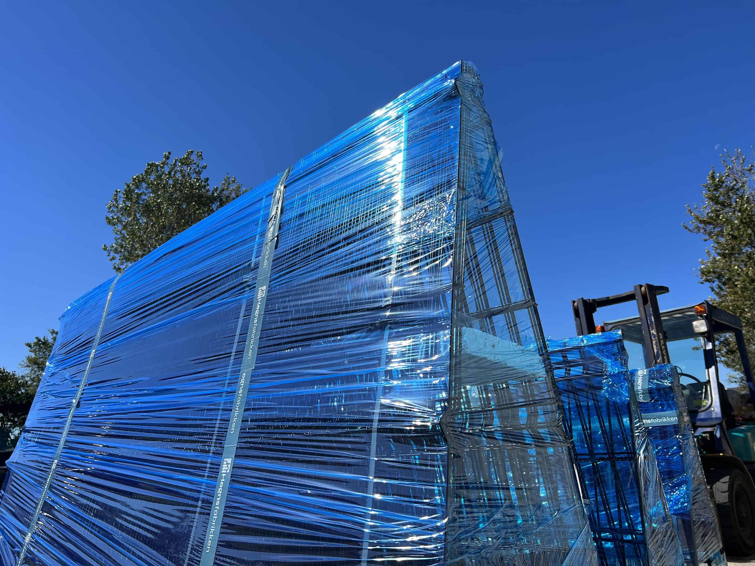 En stor stak metalrammer eller paneler pakket ind i blå plastik sidder udendørs under en klar blå himmel. En gaffeltruck er placeret ved siden af stakken i højre side, klar til levering, med træer synlige i baggrunden.