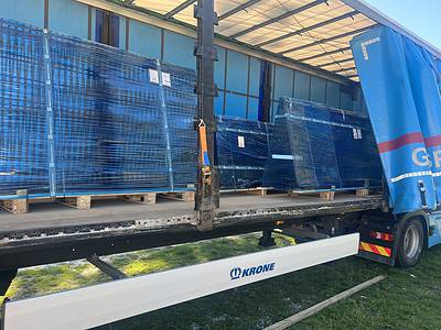 En lastbil med "KRONE" på står parkeret med sidegardinet åbent, hvilket afslører flere store blå paller forsvarligt pakket ind i plastik. Pallerne er pænt arrangeret på træbaser inde i lastbilen, klar til levering eller aflæsning.