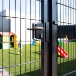En låst metalport med en Locinox overflademonteret låsekasse komplet har udsigt til en legeplads med kunstgræs, farverige legehuse, rutsjebaner og slørede børn, der leger i baggrunden.