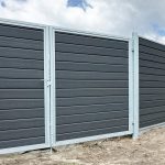 A Dobbeltlågeramme med lås - Varmgalvaniseret/Pulverlakeret - til 25mm komposit brædder har mørkegrå vandrette paneler og sølvfarvede stolper, der står på bar mark under en delvist overskyet himmel.