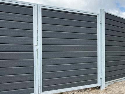 A Dobbeltlågeramme med lås - Varmgalvaniseret/Pulverlakeret - til 25mm komposit brædder har mørkegrå vandrette paneler og sølvfarvede stolper, der står på bar mark under en delvist overskyet himmel.