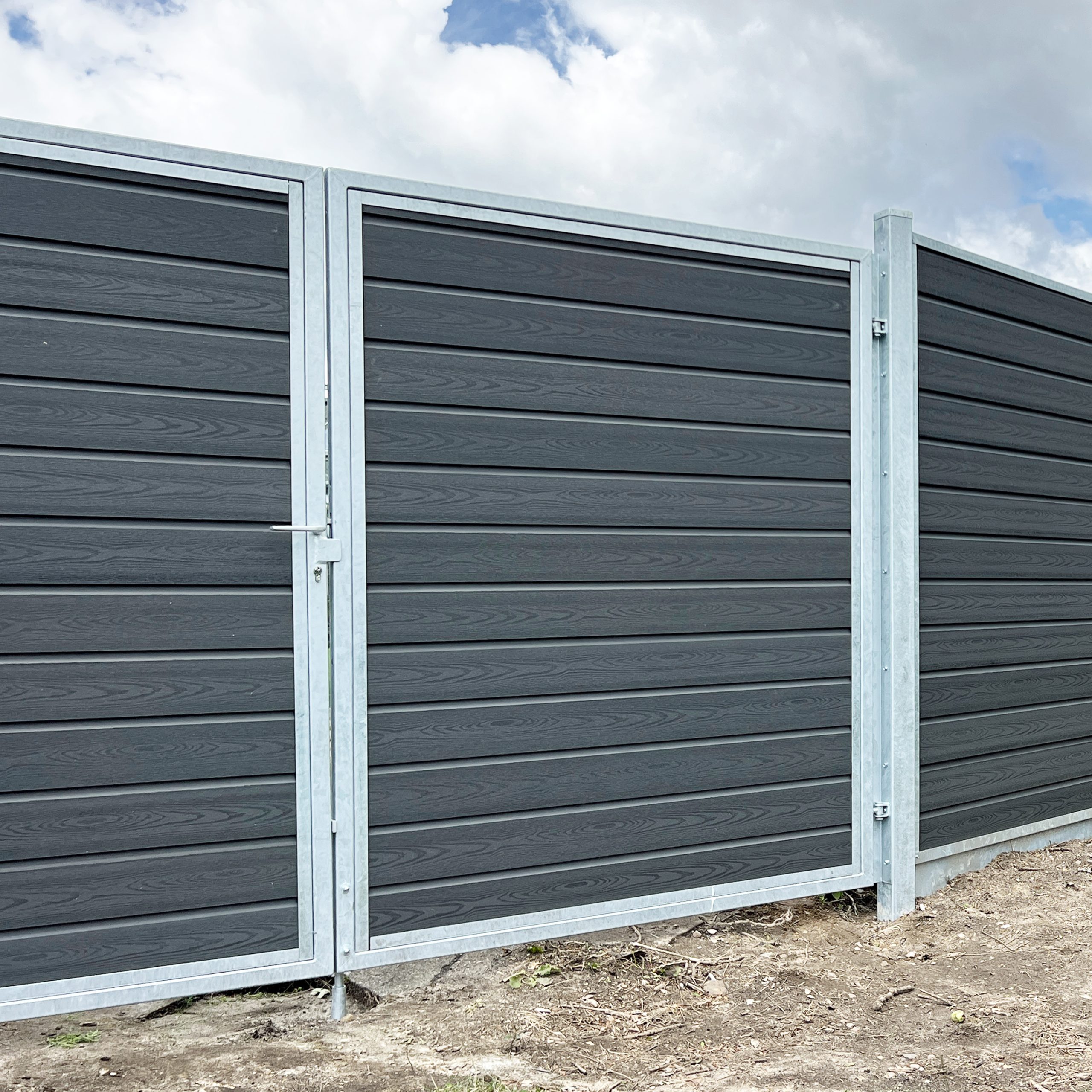 A Dobbeltlågeramme med lås - Varmgalvaniseret/Pulverlakeret - til 25mm komposit brædder har mørkegrå vandrette paneler og sølvfarvede stolper, der står på bar mark under en delvist overskyet himmel.