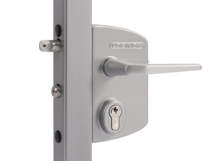 Et nærbillede af en sølv Locinox låsekasse-mekanisme installeret på en metalramme. Den overflademonterede lås har et håndtag, et nøglehul til en cylindrisk nøgle, og mærkenavnet "Locinox" er synligt på låsekroppen. En metallås strækker sig fra låsen.