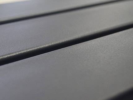 Nærbillede af en glat, mørkegrå Balusterlåge metal dobbeltdør overflade med parallelle riller, der løber på tværs. Den matte tekstur og riller kaster skygger og tilføjer dybde til billedet. Perspektivet antyder, at det kunne være en bordplade eller industripaneler.