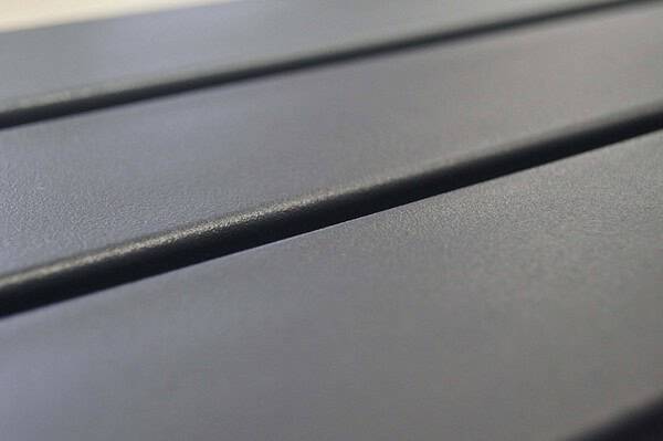 Nærbillede af en glat, mørkegrå Balusterlåge metal dobbeltdør overflade med parallelle riller, der løber på tværs. Den matte tekstur og riller kaster skygger og tilføjer dybde til billedet. Perspektivet antyder, at det kunne være en bordplade eller industripaneler.