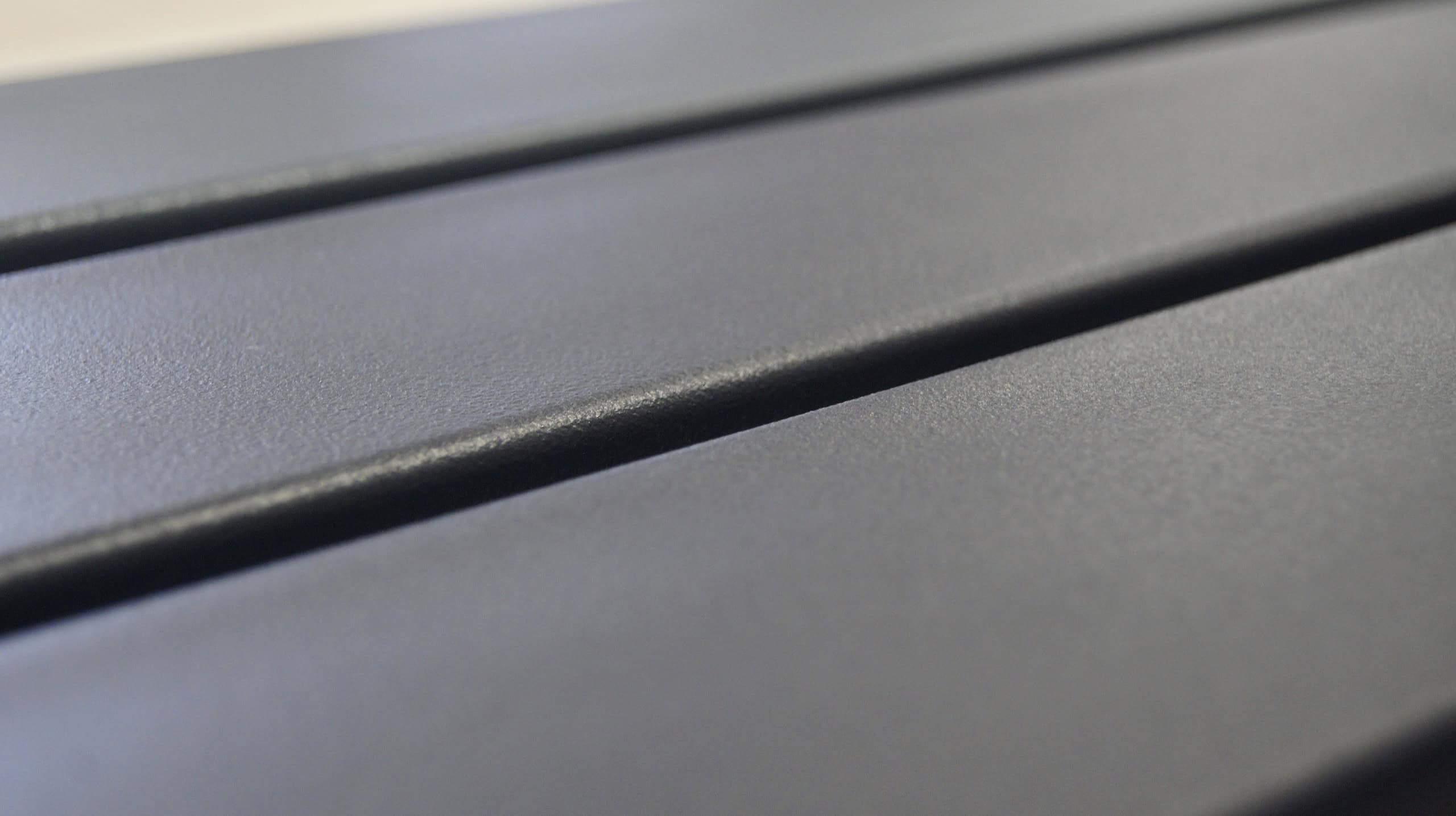 Nærbillede af en glat, mørkegrå Balusterlåge metal dobbeltdør overflade med parallelle riller, der løber på tværs. Den matte tekstur og riller kaster skygger og tilføjer dybde til billedet. Perspektivet antyder, at det kunne være en bordplade eller industripaneler.