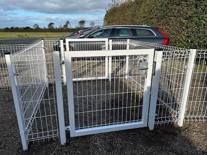 Et kompakt kabinet lavet af HF PREMIUM 3D 5/5mm Panelhegn, pulverlakeret i hvid RAL 9010, vises udenfor på en grusoverflade. Strukturen har en port med en lås og er i øjeblikket tom. I baggrunden ses flere parkerede biler og en hæk under en delvist overskyet himmel.