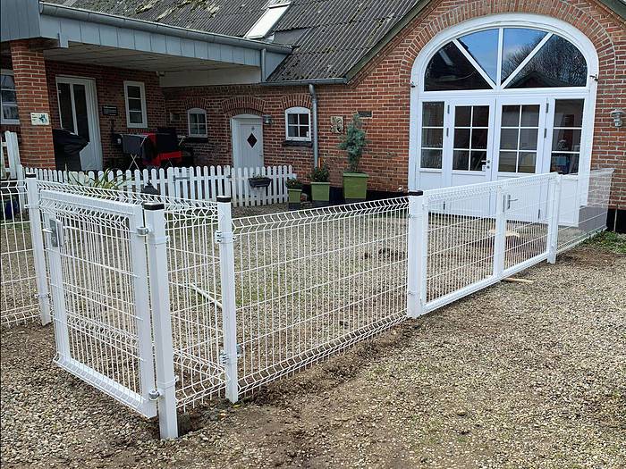 Et hvidt metalhegn, specifikt Panelhegn 3D 5/5mm - HF PREMIUM Pulverlakeret Hvid RAL 9010, omslutter et område foran et murstenshus med gråt tag. Hegnet omfatter en låge og omkranser noget grus. I baggrunden ses en større bygning med en buet, vinduesdør og et mindre hvidt stakit.