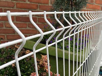 Nærbillede af en Panelhegn 3D 5/5mm - HF PREMIUM Pulverlakeret Hvid RAL 9010 med zigzag mønster foran en murstensvæg. Bag hegnet er der en grøn plantekasse fyldt med forskellige blomster og planter, blandt andet lilla blomster og grønt.