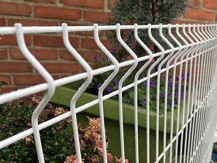 Nærbillede af en Panelhegn 3D 5/5mm - HF PREMIUM Pulverlakeret Hvid RAL 9010 med zigzag mønster foran en murstensvæg. Bag hegnet er der en grøn plantekasse fyldt med forskellige blomster og planter, blandt andet lilla blomster og grønt.