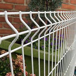 Nærbillede af en Panelhegn 3D 5/5mm - HF PREMIUM Pulverlakeret Hvid RAL 9010 med zigzag mønster foran en murstensvæg. Bag hegnet er der en grøn plantekasse fyldt med forskellige blomster og planter, blandt andet lilla blomster og grønt.