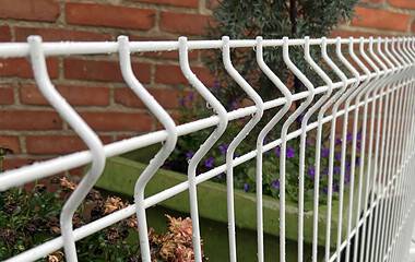 Nærbillede af en Panelhegn 3D 5/5mm - HF PREMIUM Pulverlakeret Hvid RAL 9010 med zigzag mønster foran en murstensvæg. Bag hegnet er der en grøn plantekasse fyldt med forskellige blomster og planter, blandt andet lilla blomster og grønt.