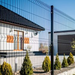 Et moderne hus med sort tegltag og træaccenter ses bag et slankt Metal Hegn 3D Panel "HF-DESIGN SERIE" inkl. stålstolper & beslag - pulverlakeret sort hegn. Gården har pænt trimmede buske langs hegnet, og himlen er klar og blå.