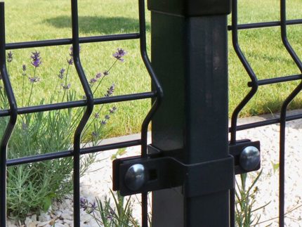 Nærbillede af et metal Hegn 3D panel "HF-DESIGN SERIE" i sort, med stålstolper og beslag, med bølgede trådnetpaneler fastgjort med bolte. Bag hegnet er grønt græs og blomstrende lavendelplanter delvist synlige sammen med små hvide sten på jorden.