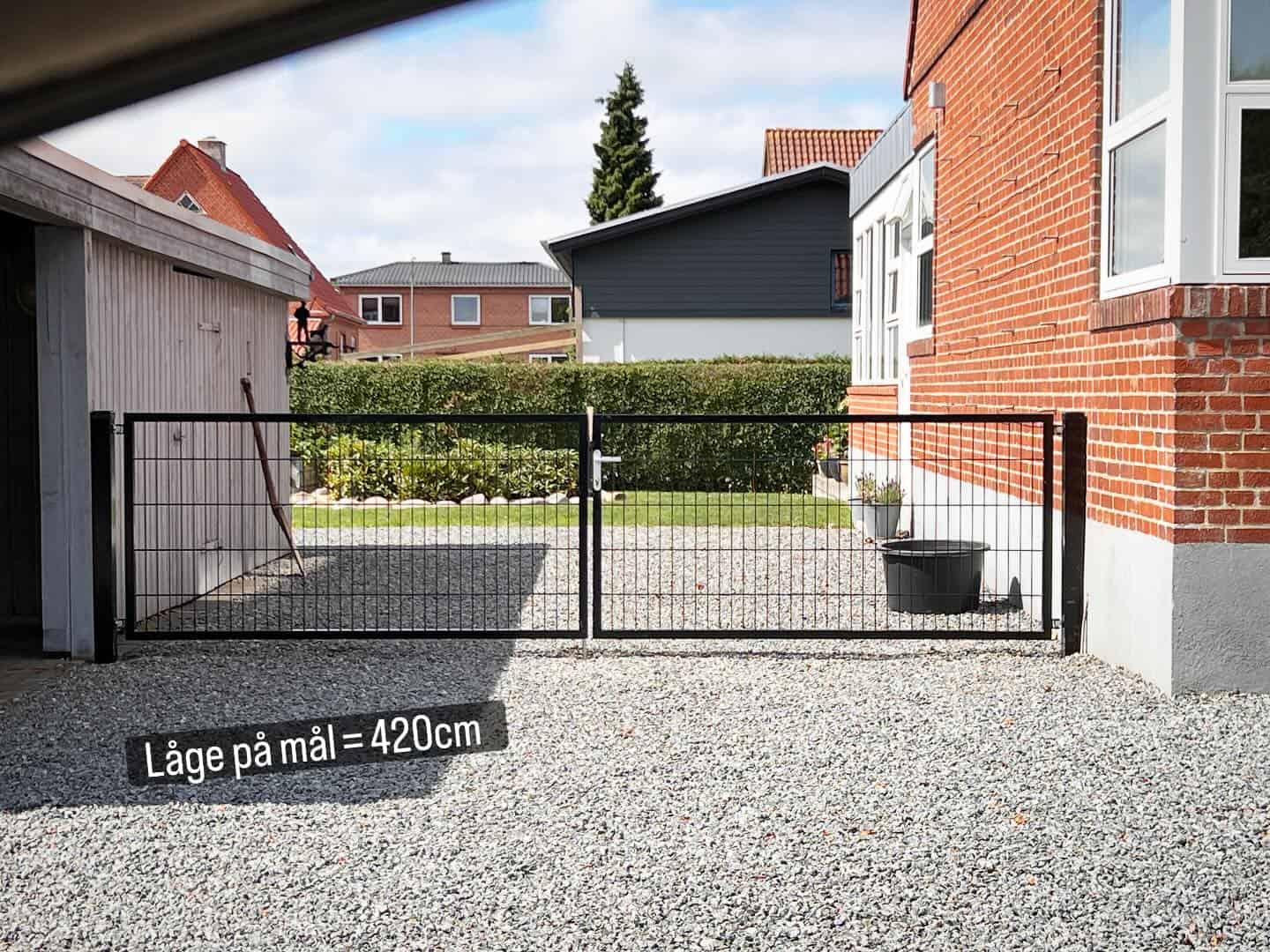 En sort metalport på 420 cm står mellem et murstenshus og en garage på en grusbelagt indkørsel med hække og nabohuse i baggrunden. En blomsterkrukke er placeret nær porten.