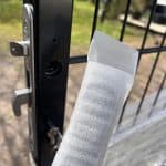 Et nærbillede af en metalport med et Dækskjold til låseindsats sæt Sort Alu, vist delvist åbent. I forgrunden holder en hånd en hvid skumbeskyttelsesliste nær porten, mens sløret grønt er synligt i baggrunden.