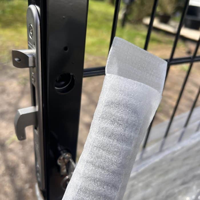 Et nærbillede af en metalport med et Dækskjold til låseindsats sæt Sort Alu, vist delvist åbent. I forgrunden holder en hånd en hvid skumbeskyttelsesliste nær porten, mens sløret grønt er synligt i baggrunden.