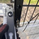 En behandsket hånd holder en sort lås ved siden af et Sort Alu-hegn og viser Dækskjold til låseindsats-sættet Sort Alu med en nøgle i låseindsatsen. Lidt plastikfolie og udendørs jord er synligt i baggrunden.