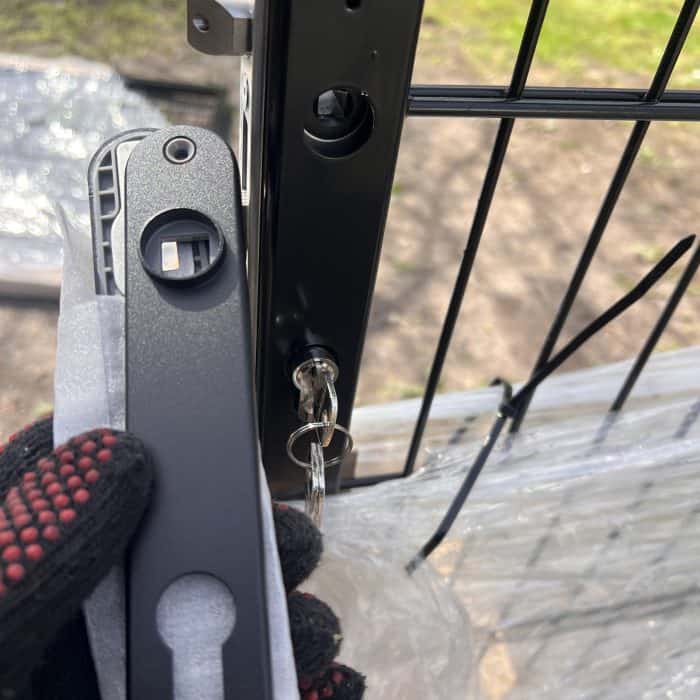 En behandsket hånd holder en sort lås ved siden af et Sort Alu-hegn og viser Dækskjold til låseindsats-sættet Sort Alu med en nøgle i låseindsatsen. Lidt plastikfolie og udendørs jord er synligt i baggrunden.