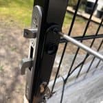 Et nærbillede af en person, der installerer et Dækskjold til låseindsats sæt Sort Alu på en sort metalport udendørs, med græs, en terrasse og den sorte alu-belægning synlig i baggrunden.