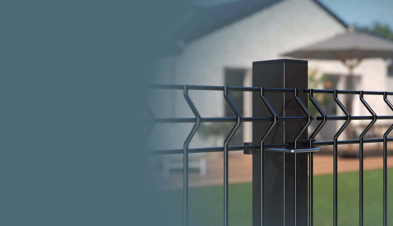 Nærbillede af et sort metalhegn med lodrette og vandrette stænger, installeret udendørs. I baggrunden er der et sløret billede af et hvidt hus med et skrånende tag og en gårdhaveparaply, hvilket tyder på et beboelsesmiljø. Den galvaniserede hegn og låger i venstre side af billedet falmer til en blå farve.